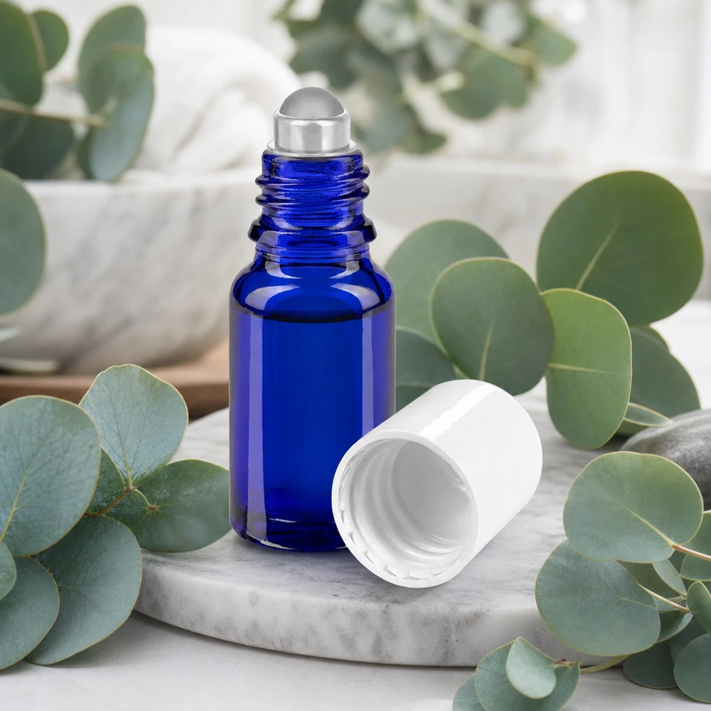 Flacon en verre bleu à bille de 10 ml, ouvert sur un plateau en marbre, entouré de branches d'eucalyptus fraîches