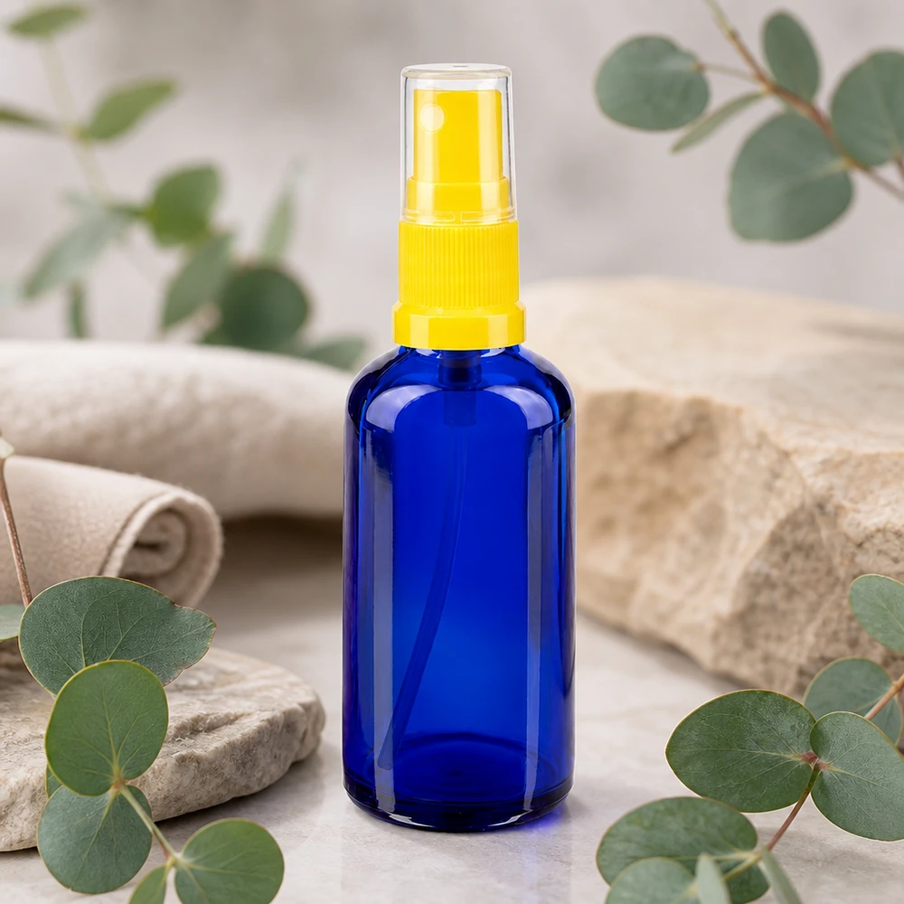 Flacon de pharmacie bleu 50ml embout spray jaune Flacon en verre bleu de 50 ml muni d'un vaporisateur STD jaune, posé entre des branches d'eucalyptus et une pierre naturelle