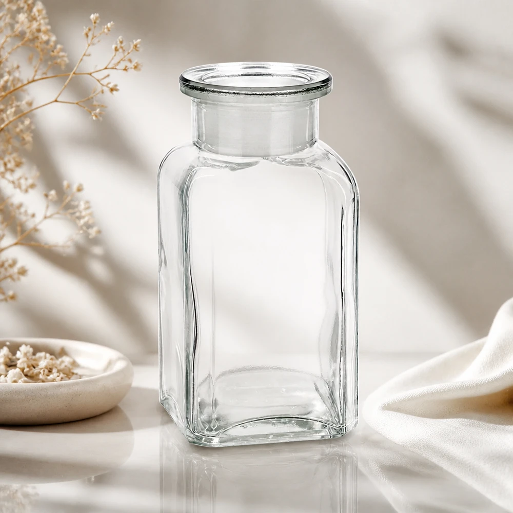 Flacon carré col large 100 ml + bouchon RIN, verre clair Petit flacon de pharmacie en verre transparent de 100 ml, de forme carrée, muni d'un bouchon, posé à côté d'un bol en céramique