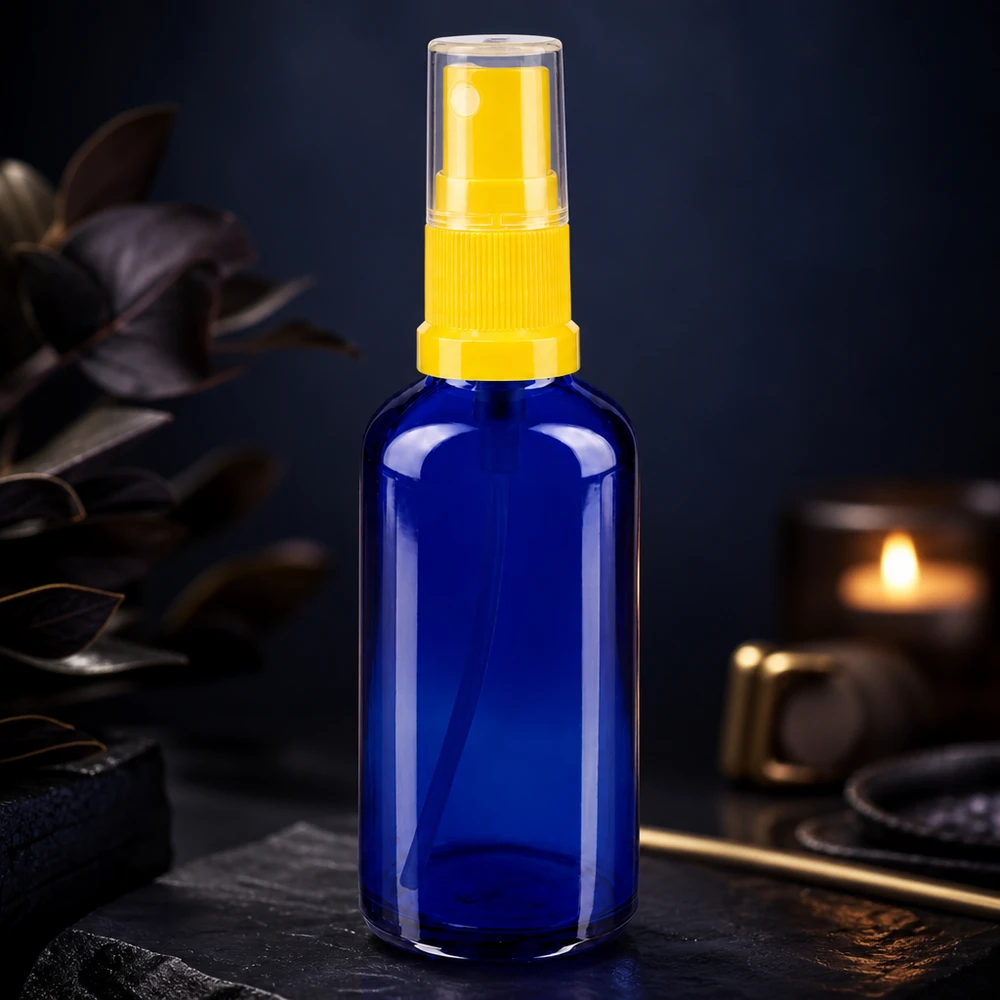 Bouteille en verre bleue de 50 ml avec un vaporisateur jaune, à côté d'une bougie et de feuilles sombres