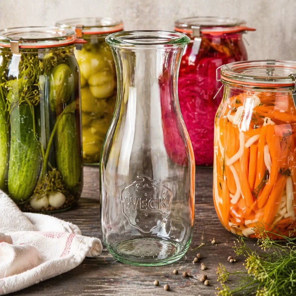 Bouteille de jus WECK RR60 de 530 ml entourée de bocaux de légumes colorés et d'accessoires de conservation sur un fond en bois.