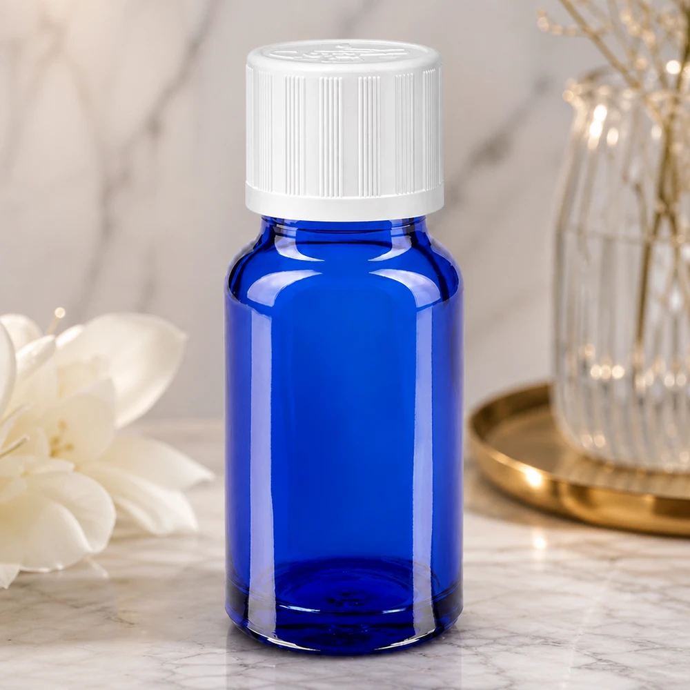 Flacon en verre bleu de 10 ml avec un bouchon à vis blanc, à côté d'une fleur blanche et d'un vase en verre