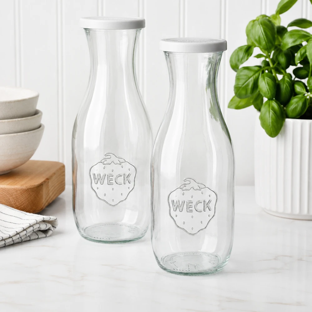 Bouteille à jus de 1 062 ml avec couvercle fraîcheur WECK RR60 Deux bouteilles de jus WECK 1062ml avec couvercle blanc dans une cuisine moderne à côté de coupes et d'une plante de basilic