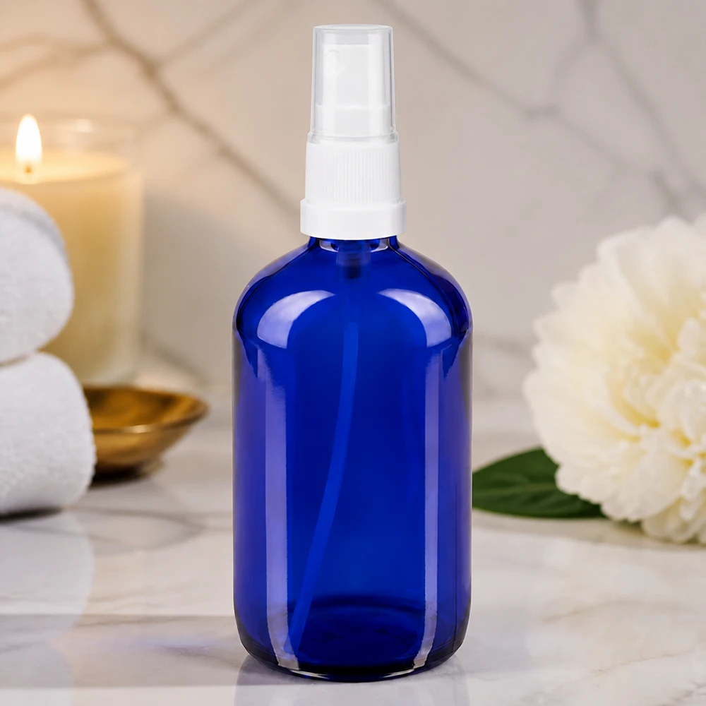 Flacon en verre bleu cobalt de 100 ml avec vaporisateur blanc, posé sur du marbre dans une ambiance spa