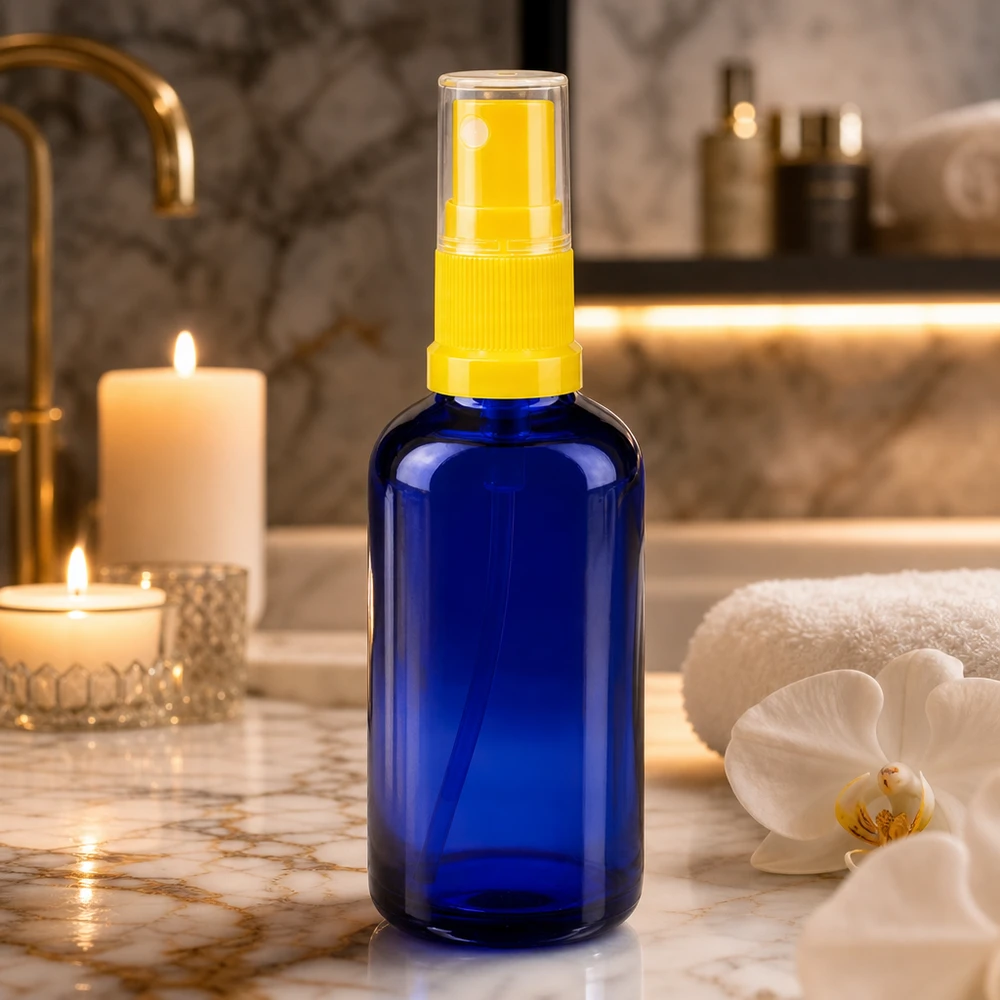 Flacon en verre bleu cobalt de 50 ml avec un vaporisateur jaune, à côté d'une orchidée blanche et de bougies